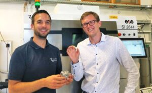 Alex Hilgenberg and Thomas Hilger holding 3D-printed parts in front of a metal 3D printer during the launch of their ceramic-metal hybrid project.